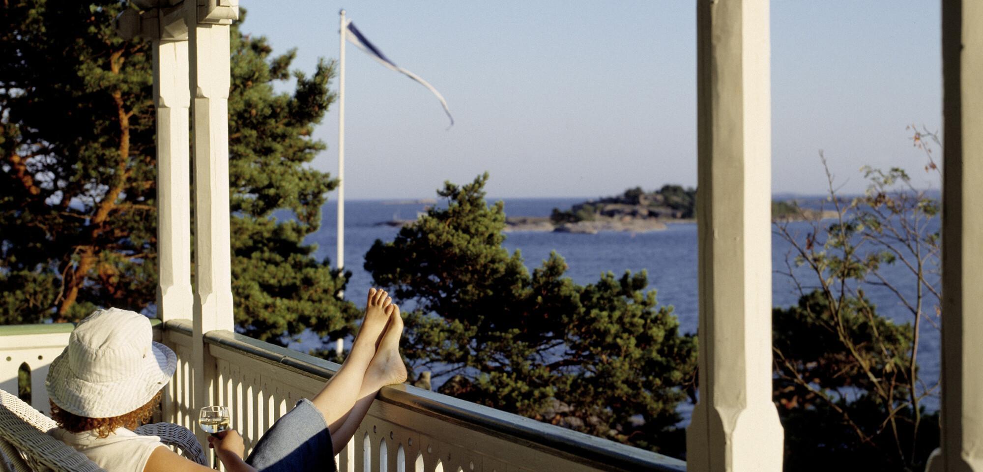 Nainen istuu pitsihuvilan parvekkeella ja katsoo merta päin. En kvinna sitter på en spetsvillas terass och tittar mot havet. A woman is sitting on the terrace of a wooden villa.