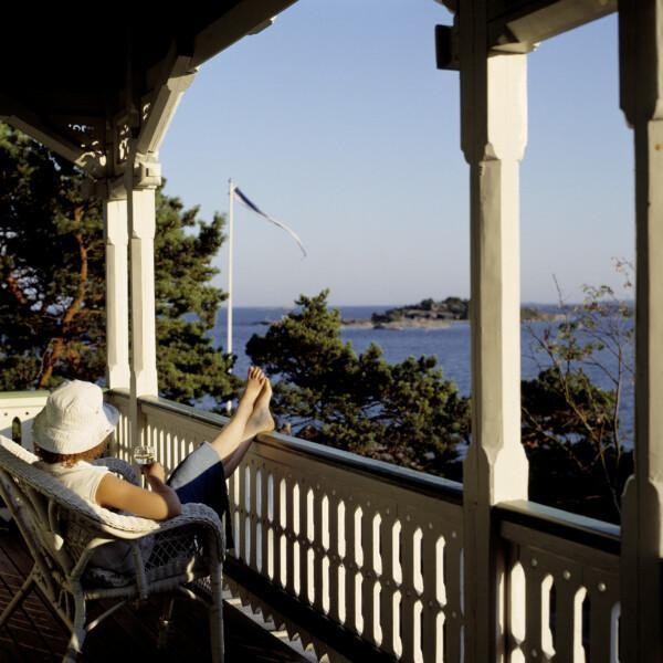 Nainen istuu pitsihuvilan parvekkeella ja katsoo merta päin. En kvinna sitter på en spetsvillas terass och tittar mot havet. A woman is sitting on the terrace of a wooden villa.